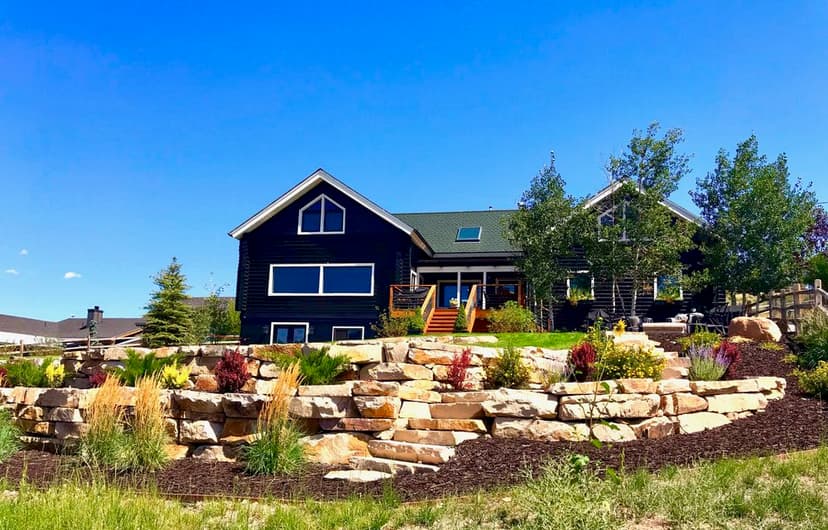 Modern black house with stone landscaping and vibrant greenery under a blue sky.
